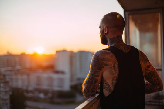tattooed guy standing on balcony overlooking city at sunrise, aspirational mood, warm soft light, cinematic framing.