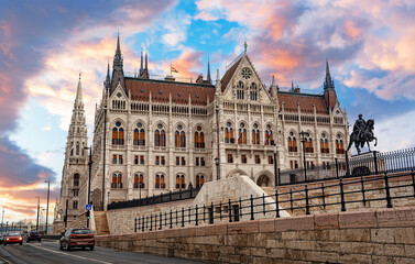 Naklejka premium The Hungarian Parliament Building stands under a dramatic sunset sky in Budapest, featuring its intricate Gothic Revival facade and a statue on the embankment.