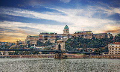 Fototapeta premium A panoramic view of the historic Buda Castle and the Szechenyi Chain Bridge spanning the Danube River under a dramatic sunset sky in Budapest, Hungary.