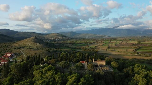 Scenic aerial view of the Necromanteion ruins surrounded by lush greenery and the expansive Acheron Valley, with mountains in the background under a partly cloudy sky.