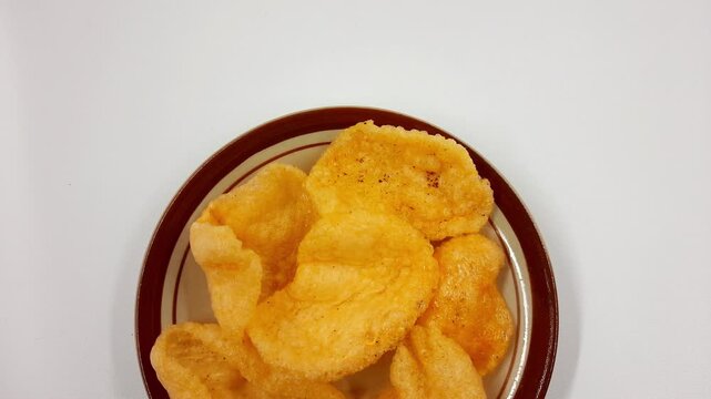 Hand slightly moves a small plate filled with golden orange fried prawn crackers against a bright white background surface in this footage.
