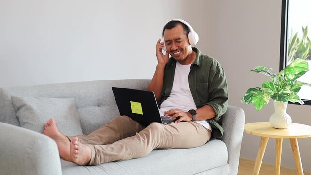 An Asian composer man sitting comfortably on a grey sofa at home, wearing white headphones and using a laptop, editing a song