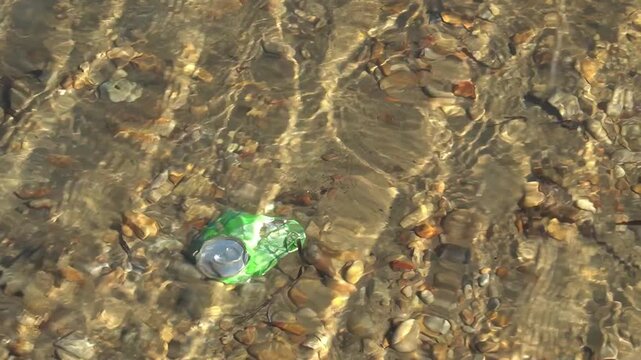 The clear water of the lake with a visible bottom, among the stones lies a green plastic bottle as an example of pollution of nature.