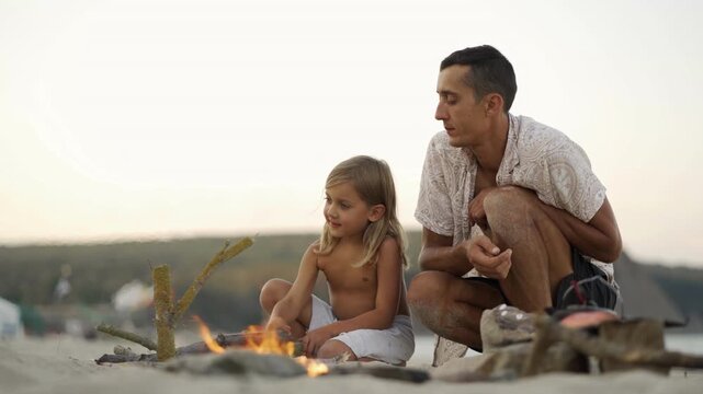 A young girl and her father relax by a campfire on the beach, sharing a quiet bonding moment at sunset. The serene atmosphere highlights the beauty of outdoor family adventures and summer vacations