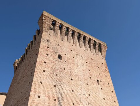 Torre medievale in mattoni chiari con merlature e machicolazioni, tipica dell&rsquo;architettura difensiva italiana, ripresa contro un cielo limpido