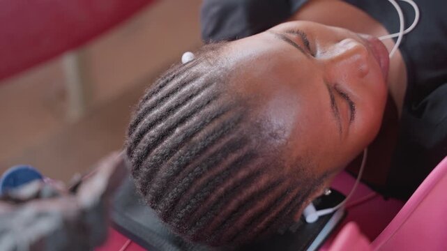 black woman athlete with cornrows listening to music during shampoo rinse at basin, glossy wet scalp and neat braid rows, pink salon chair background and attentive stylist creating peaceful care