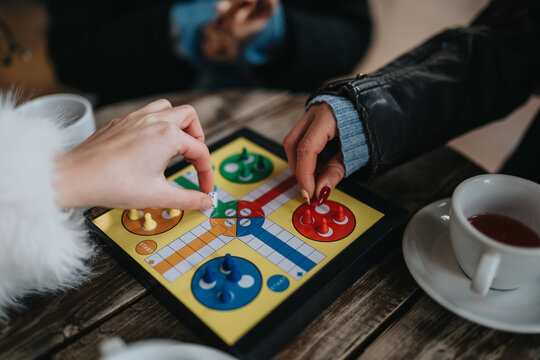 Two girls play a colorful Ludo game at a cafe table beside a cup of tea.