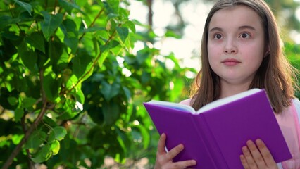 Fototapeta premium Child with backpack on back rushes to school. Teenage girl walks down street reading textbook. Student rushes to school for lesson, reading book in park. Children's education concept . Schoolchild