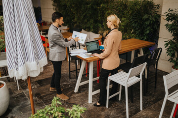 Fototapeta premium Two professionals discuss a chart in an outdoor cafe setting with a laptop. They review documents and collaborate on a project at a casual business meeting.