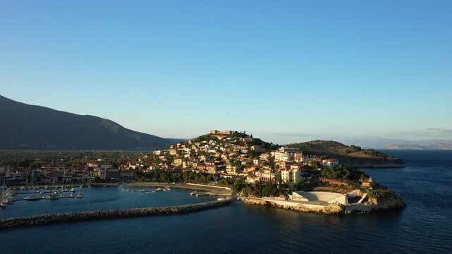 Aerial view of the picturesque seaside town of Paralio Astros with its marina, hillside houses, and clear blue waters under a bright sky. The tranquil harbor and Mediterranean landscape create a