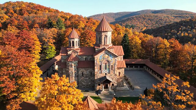 Aerial drone footage captures a historic monastery nestled amidst a vibrant forest ablaze with autumn foliage during golden hour.