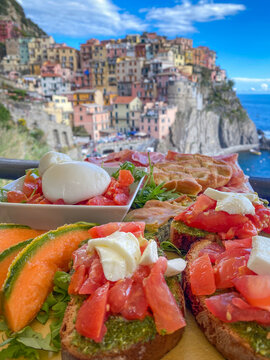 Mixed bruschetta platter with pesto, tomato, mozzarella, burrata, melon and focaccia on a terrace with colorful houses of Manarola blurred in the background, Cinque Terre, Liguria, Italy