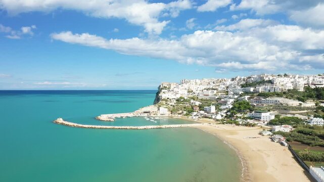 Scenic aerial view of Peschici with turquoise sea, sandy beach, and white hillside buildings under a blue sky with clouds. Picturesque coastal town in southern Italy.