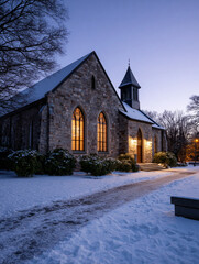 Traditional Stone Church Exterior at Winter Dusk with Glowing Stained Glass Windows and Snow Covered Holly Bushes