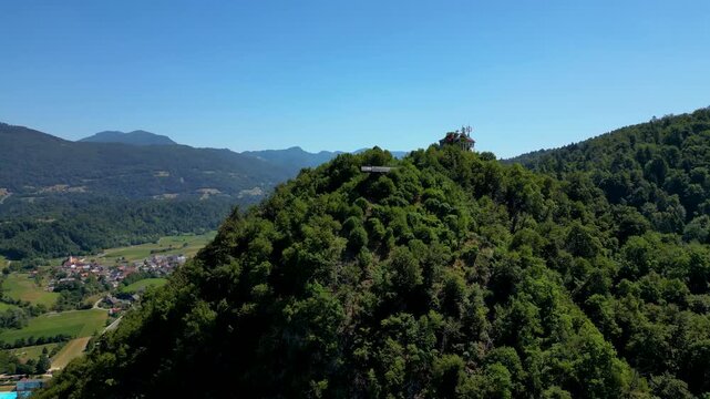 Aerial view of the lush, tree-covered hill of Stari Grad (Old Castle) in Kamnik, Slovenia, featuring a modern viewing platform and mountain hut overlooking the scenic Kamnik-Savinja Alps.