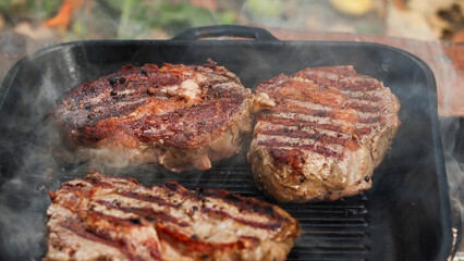 Three juicy ribeye steaks sizzling and smoking on a cast iron grill pan outdoors