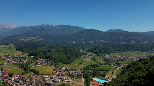 Wide aerial view of the historic town of Kamnik, Slovenia, featuring red-roofed houses, a central church, and a sports stadium, set against the towering peaks of the Kamnik-Savinja Alps in summer.