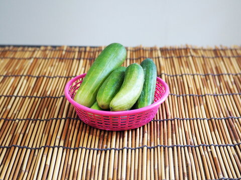 Organic cucumbers, harvested from the backyard garden, are placed in a pink plastic basket on a bamboo mat. They are popular vegetable for eat fresh fruit, cooking or decorate food plate. Cucurbitacea