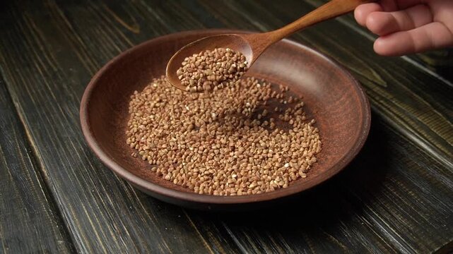A hand holds a wooden spoon pouring buckwheat seeds into a brown bowl on a dark wooden table, demonstrating a gradual movement of the action
