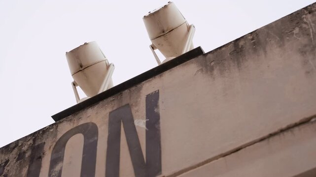 Rooftop water tanks against pale sky, aged beige containers perched on concrete parapet with bold painted letters visible, soft overcast light highlighting textured grime and rust, minimal urban