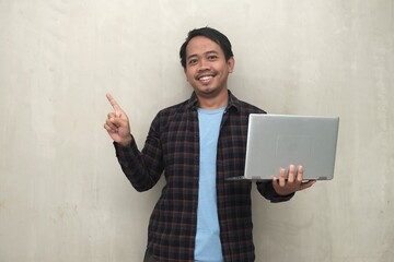 Southeast Asian man smiles while holding a laptop and presenting with open hand against plain wall, ideal for promo copy space.