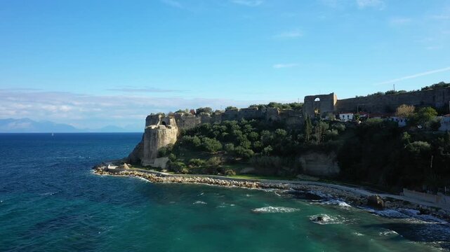 A scenic view of ancient fortress ruins overlooking the turquoise sea in Koroni, Greece, with lush greenery and dramatic cliffs under a clear sky.