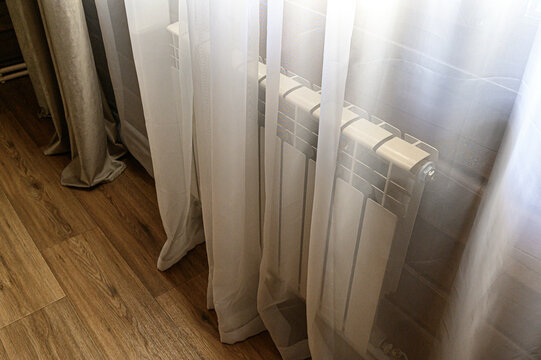 A white bimetallic radiator partially covered by sheer milk-colored curtains. Set against a wooden floor, soft light filters through the delicate fabric