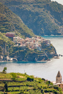 Scenic view of Manarola on the Cinque Terre coast with Corniglia bell tower in the foreground, colorful house on cliffs, and calm sea under a hazy sky in Italy