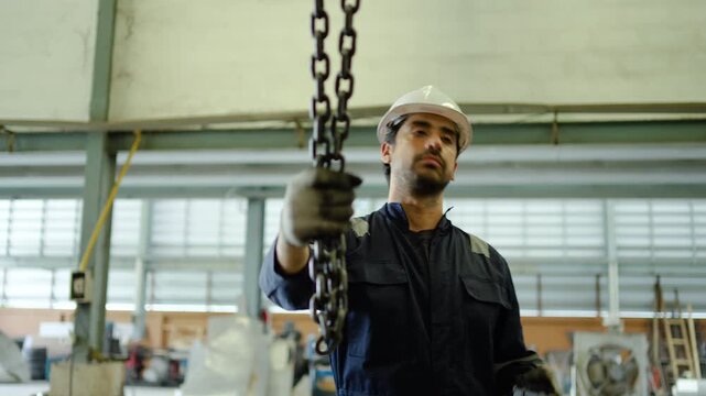 Professional male industrial technician in safety gear operates a heavy duty chain hoist and crane remote control inside a large manufacturing factory or modern warehouse facility.