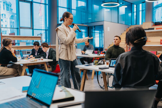 A woman speaks into a microphone in a bright coworking space while colleagues listen. The scene shows a collaborative work environment with laptops and casual seating.