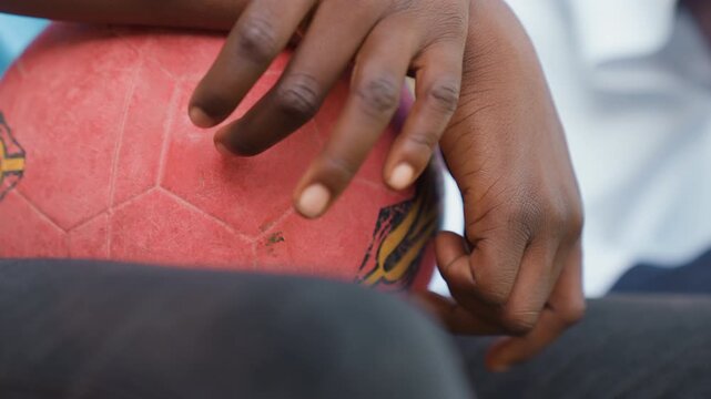 Closeup hands on red ball, school player adjusting grip with subtle scar on finger, candid team moment capturing worn emblem and sandy texture, focus on determination and resilience