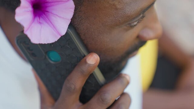 Black man closeup holding phone with purple flower, focused ear and finger on case, textured handset detail, sweat on skin, intimate private conversation, soft background blur, concentrated