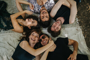 A group of six friends lies in a circle on a blanket outdoors, smiling and laughing. They relax...