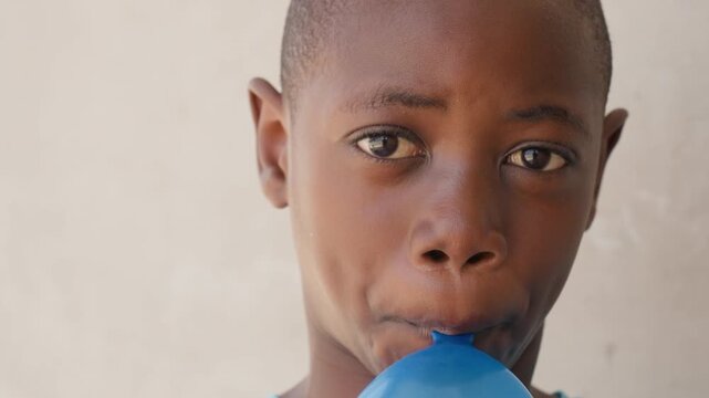 Child inflating blue balloon closeup, concentrated expression with pursed lips and sandy hands gripping balloon, plain wall background, intimate portrait of preparation for neighborhood party, soft