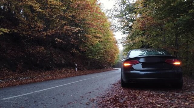 Broken black passenger car with engine failure and emergency flashing light is parked on side of mountain rural road in forest waiting for tow service