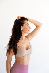 Fototapeta premium Brunette 30s caucasian Woman looking away in sports bra and leggings posing with arms up holding hair in hands in daylight studio isolated on white background. Sexy fit female body in Workout Clothes.