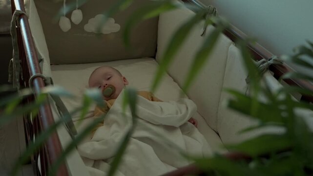 Infant sleep in cozy baby crib with minimalist bedding. white baby with pacifier swaddled in soft blanket covering tiny body. Dim nursery light. Relaxed arms and slow breathing. selective focus 