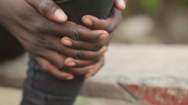 Black athlete pressing knee with both hands on outdoor bench, closeup of fingers and fabric showing pressure and tenderness, intimate portrayal of postimpact pain and search for relief, suitable