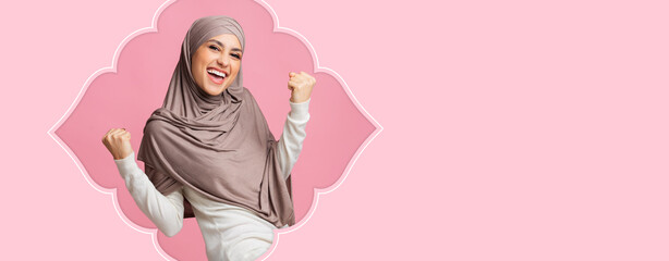 A woman wearing a hijab shows excitement with her arms raised. She stands against a pink background. Her expression conveys happiness and energy. © Prostock-studio