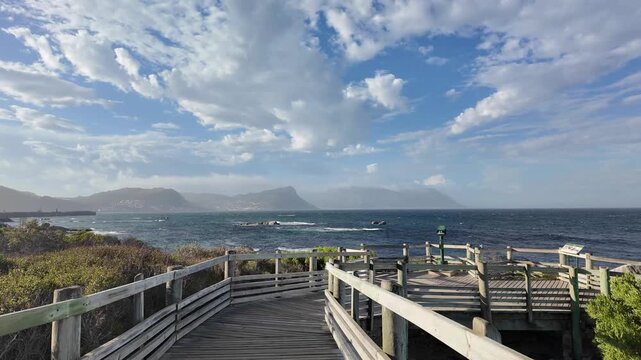 Simon's Town South Africa. 06.02.2026. Video. A wooden walkway with railings leads to a viewpoint offering expansive ocean vistas and hazy mountain range of Cape Peninsular under a dramatic sky