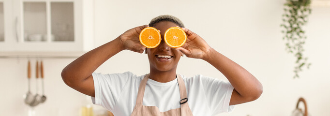 A person stands in a bright kitchen holding two orange slices in front of their face. They smile happily. The kitchen has modern decor and plants in the background, creating a lively atmosphere. © Prostock-studio