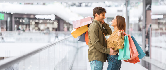 A couple stands close to each other in a mall, smiling and holding shopping bags. They are surrounded by a busy shopping environment with various stores in the background. © Prostock-studio