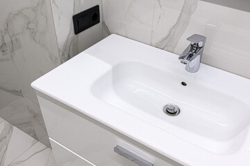 Fototapeta premium A sleek white ceramic sink with a chrome faucet is mounted on a white vanity cabinet; set against marble-patterned tiles with a black wall outlet nearby.