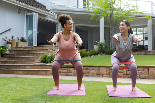 Diverse female friends squatting with loop bands on pink mats in backyard wearing workout clothes