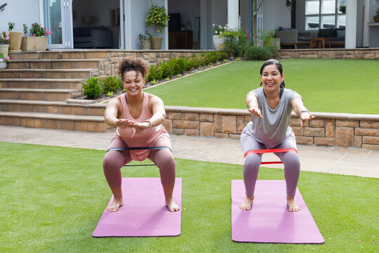Diverse female friends exercising in backyard on pink mats wearing athletic tops, using loop bands