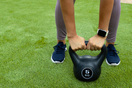 Female gripping 5 KG kettlebell while standing on lawn, wearing lilac leggings and white smartwatch