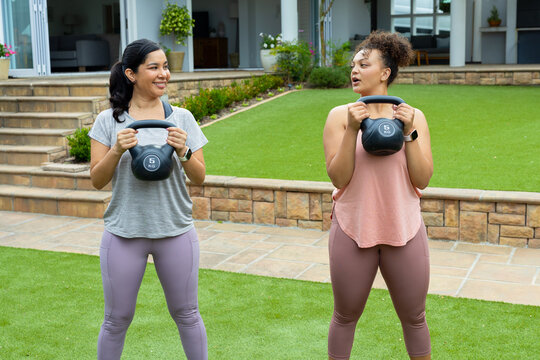Diverse female friends holding 5kg kettlebells in sportswear and wearing smartwatches in backyard
