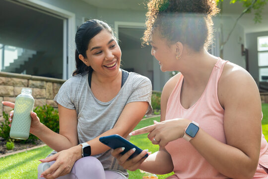 Diverse female friends sitting on front lawn in athletic wear, holding smartphone and water bottle