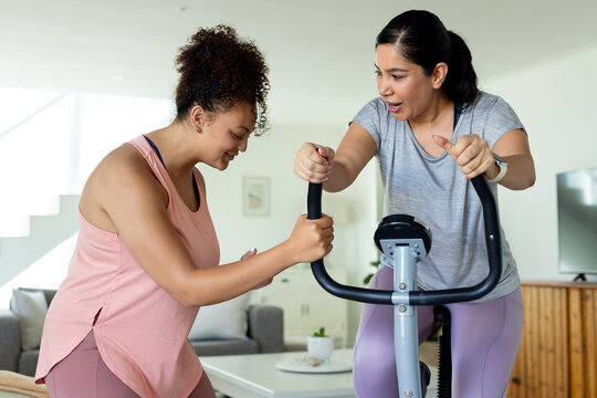 Diverse female friends in sportswear pedaling bike at home, one supporting rider wearing smartwatch