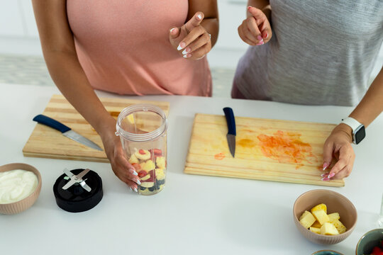 Diverse female friends standing at kitchen counter preparing fruit with clear blender cup